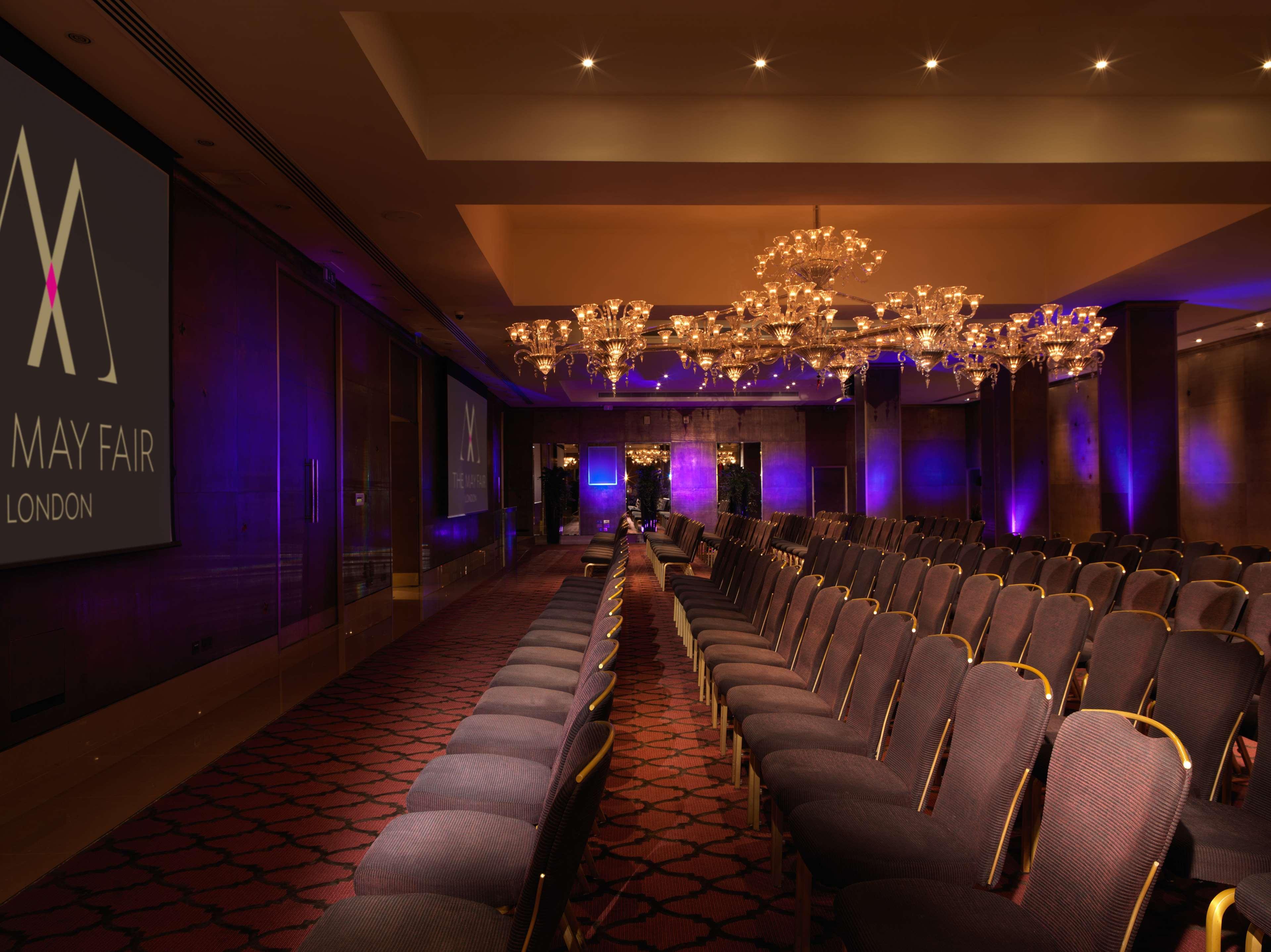 The May Fair, A Radisson Collection Hotel, Mayfair London Exterior photo The image shows a conference or event space with rows of elegant, upholstered chairs facing a large screen. The room is adorned with chandeliers that provide ambient lighting, and there are purple lighting accents around the space. The branding for "