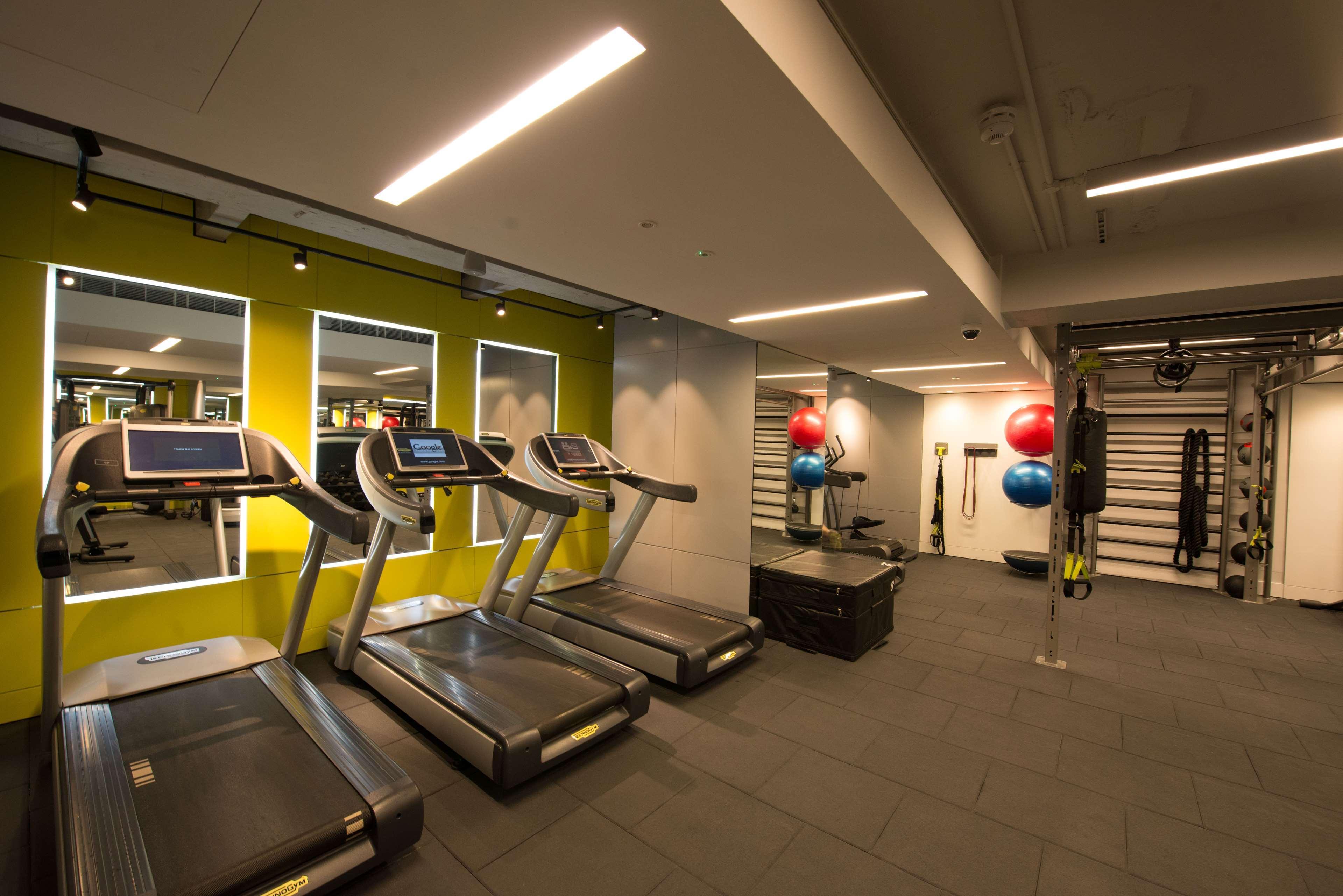 The May Fair, A Radisson Collection Hotel, Mayfair London Exterior photo The photo shows a modern gym interior featuring several exercise equipment. In the foreground, there are three treadmills aligned along a wall with mirrors. The wall behind the treadmills is painted a vibrant green, enhancing the energetic atmosphere