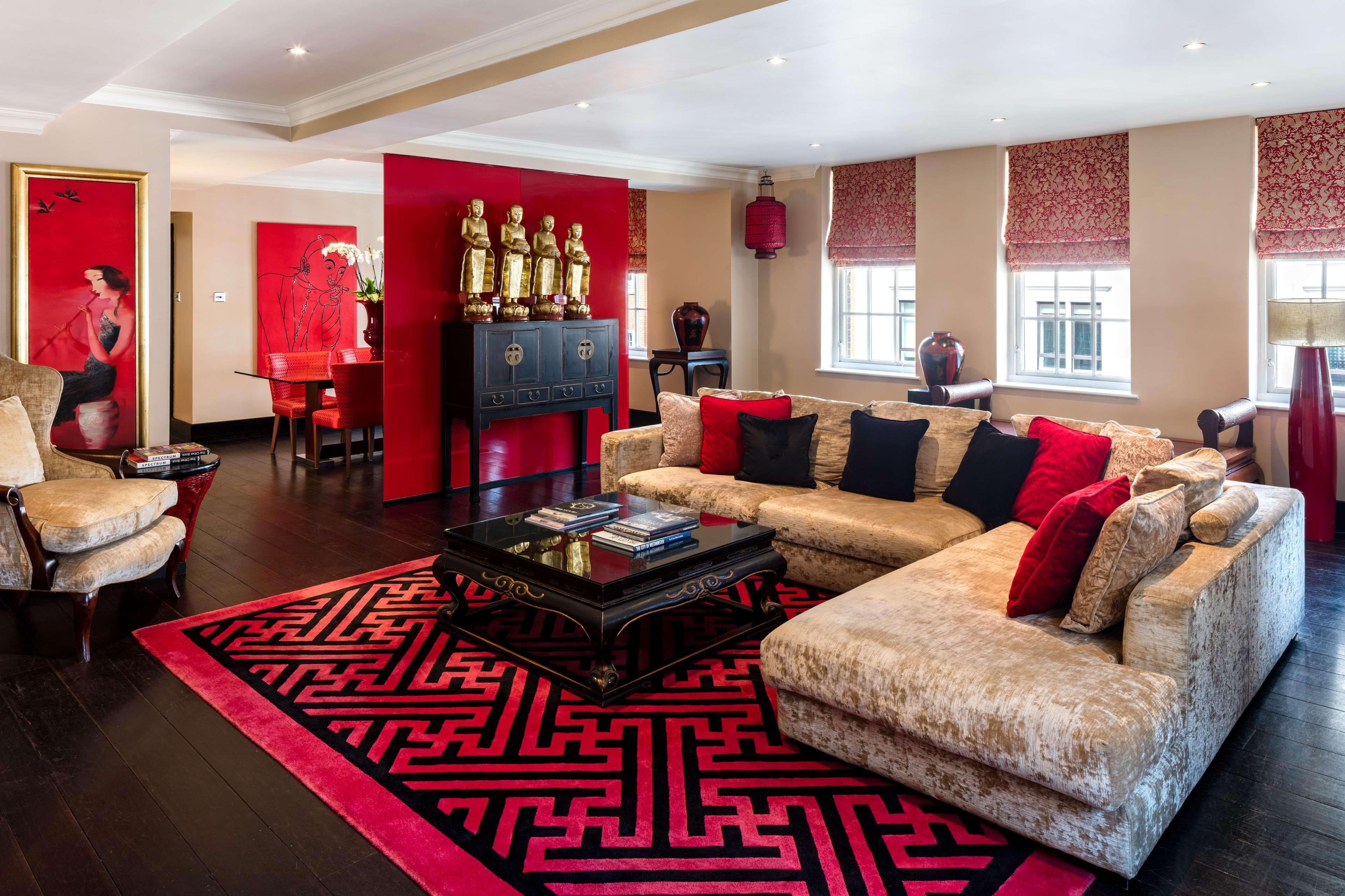 The May Fair, A Radisson Collection Hotel, Mayfair London Exterior photo The photo shows a stylish and elegant living room designed with a modern aesthetic. The space features a large, plush sofa in a light color, complemented by red and black throw pillows. A dark coffee table sits in the center, adorned with books and d