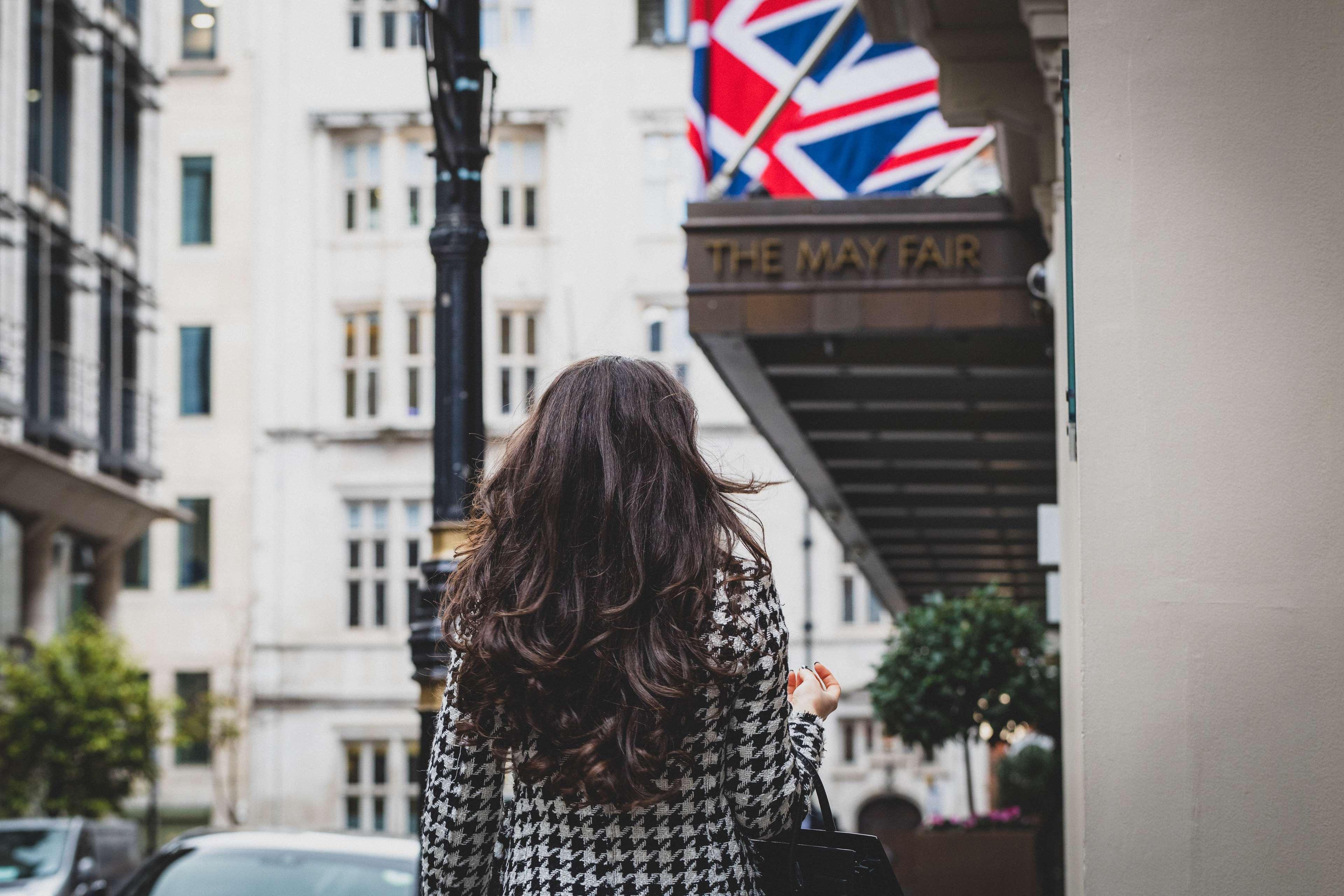 The May Fair, A Radisson Collection Hotel, Mayfair London Exterior photo The photo shows a woman with long, wavy hair standing with her back to the camera. She is wearing a houndstooth-patterned coat. In the background, there is a building with a sign that reads "THE MAY FAIR" and a British flag hanging prominently. The s