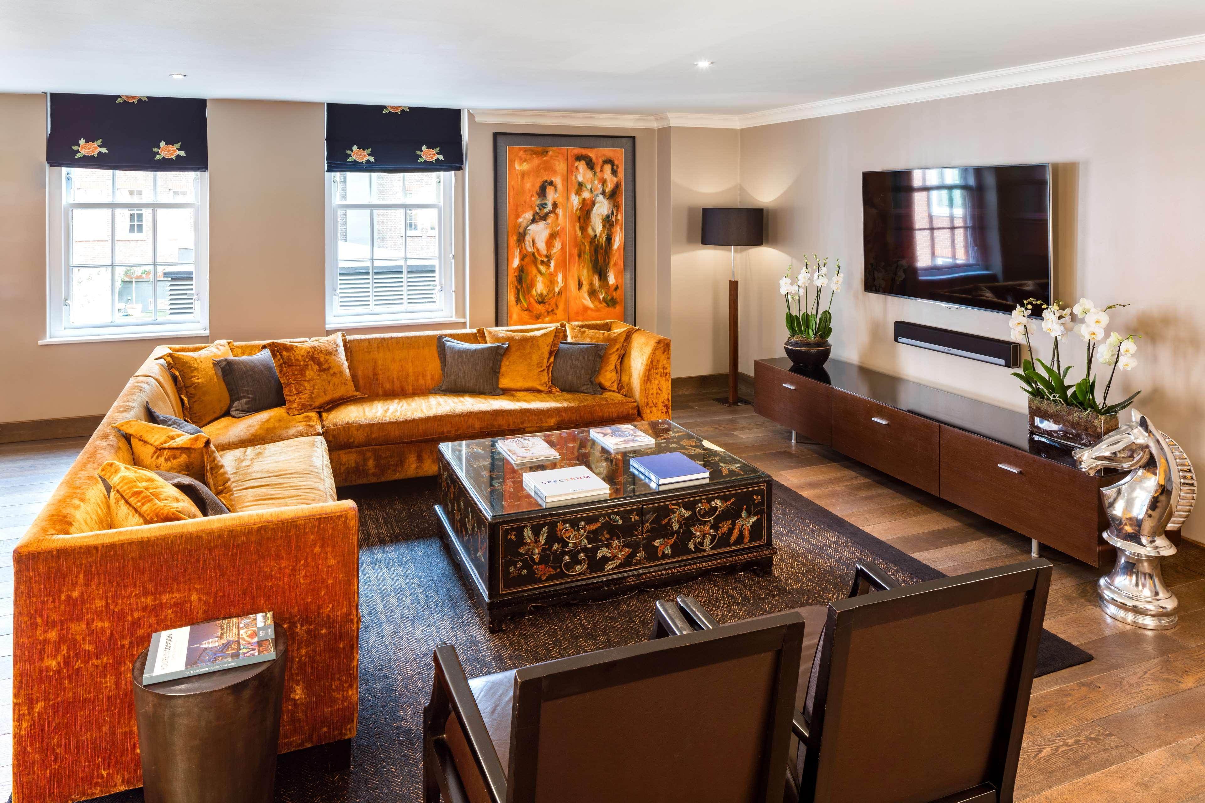 The May Fair, A Radisson Collection Hotel, Mayfair London Exterior photo The photo shows a modern living room with a stylish design. It features a large, orange sectional sofa with colorful cushions, positioned around a coffee table that has an ornate design. The floor is wooden, and there's a dark area rug beneath the se