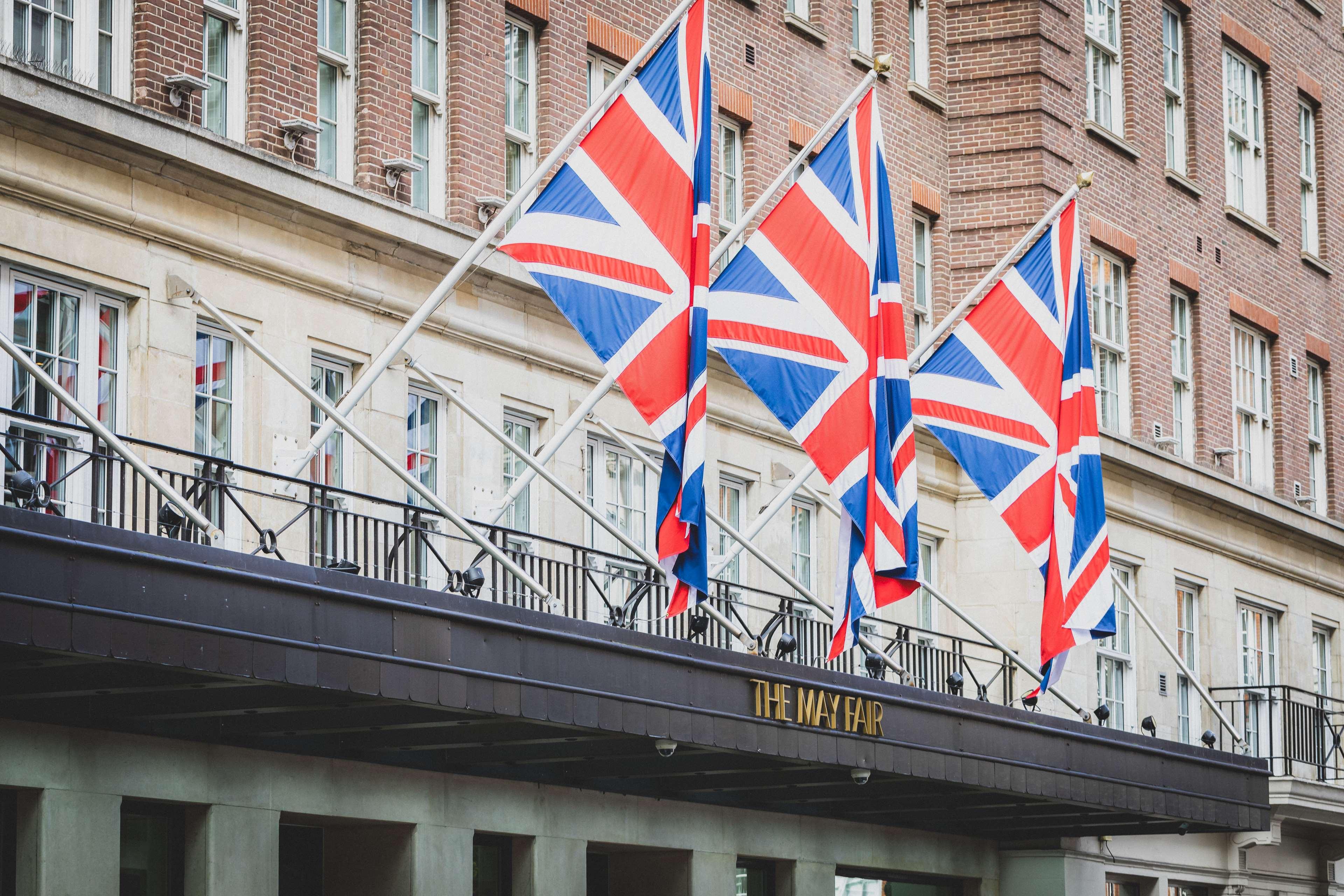 The May Fair, A Radisson Collection Hotel, Mayfair London Exterior photo The photo shows a building façade with several Union Jack flags prominently displayed. These flags are arranged in a row, indicating a British theme. The entrance of the building features a sign with the name "The Mayfair," suggesting that this could