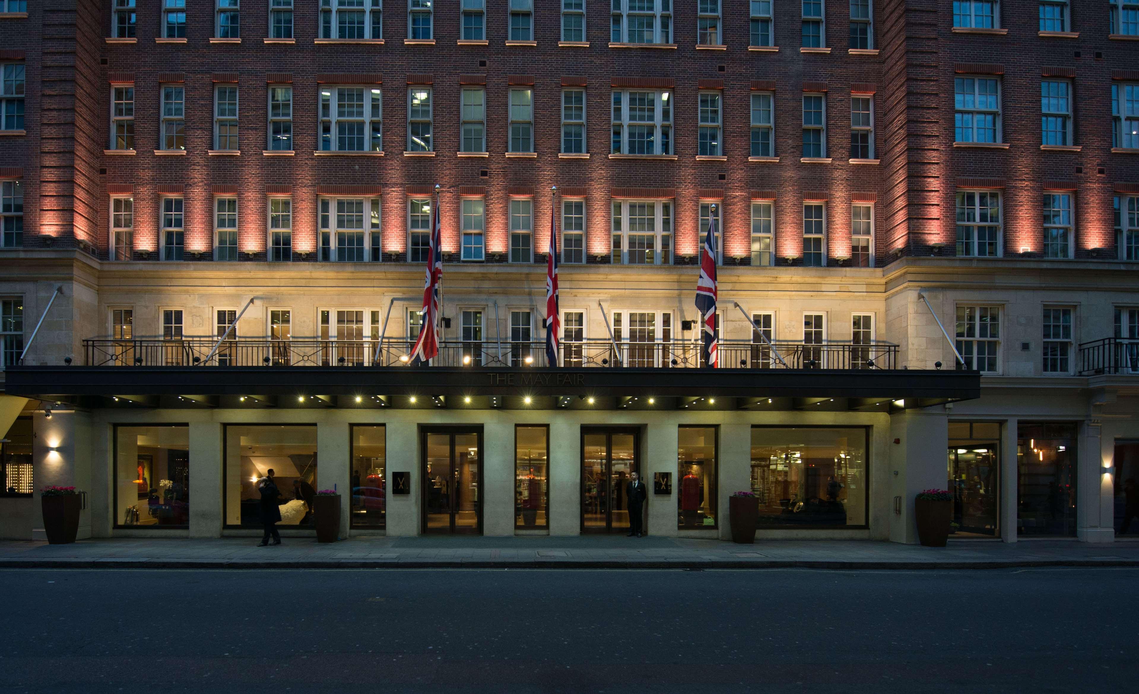 The May Fair, A Radisson Collection Hotel, Mayfair London Exterior photo The photo depicts a grand hotel exterior at dusk. The building has a classic architectural style, characterized by multiple stories with large windows. The facade is illuminated, highlighting the architectural features. British flags are prominently 