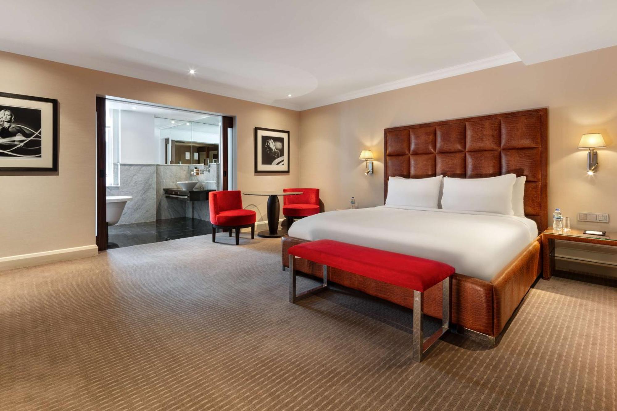 The May Fair, A Radisson Collection Hotel, Mayfair London Exterior photo The photo shows a modern hotel room featuring a large, comfortable king-size bed with a beige and brown color scheme. There are two red accent chairs positioned near the bed, adding a pop of color. A small red bench is placed at the foot of the bed. 