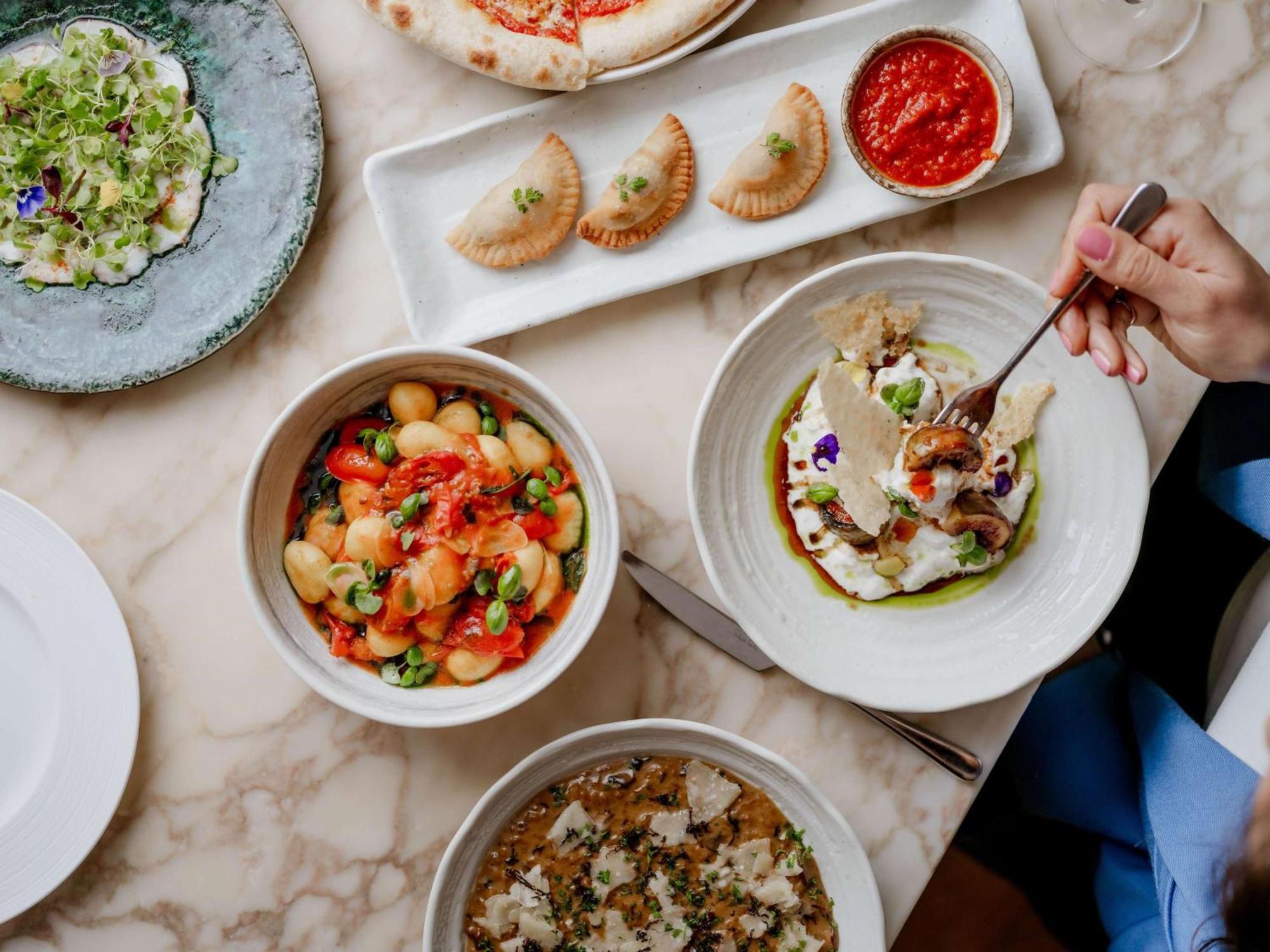 The May Fair, A Radisson Collection Hotel, Mayfair London Exterior photo The photo showcases a beautifully arranged table filled with various dishes. In the foreground, there are three bowls: one containing a colorful mix of gnocchi with vibrant toppings, another featuring a dish with what appears to be fish or seafood, g