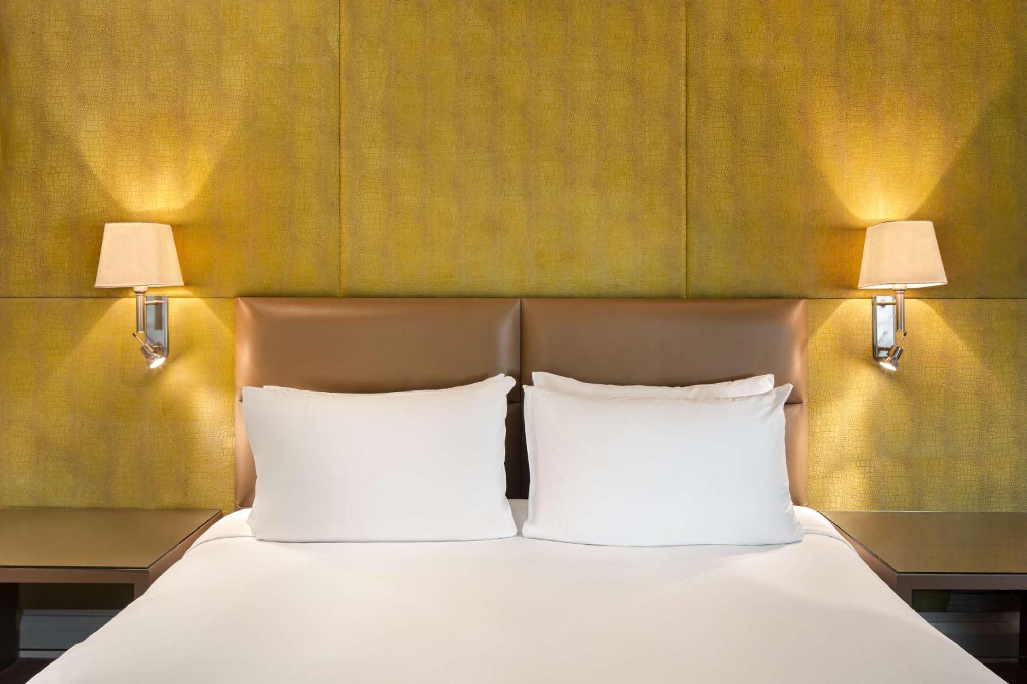 The May Fair, A Radisson Collection Hotel, Mayfair London Exterior photo The photo shows a cozy bedroom setting featuring a large bed with two white pillows. The headboard has a smooth, light brown leather material, and there are two wall-mounted lamps on either side of the bed, illuminating a textured yellow wall behind 